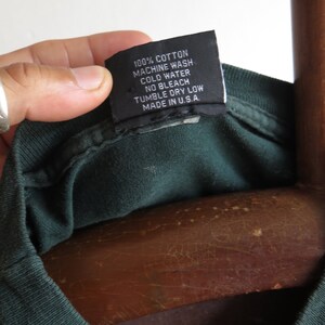 May include: A dark green t-shirt with a black care label that reads "100% Cotton, Machine Wash Cold Water, No Bleach, Tumble Dry Low, Made in U.S.A."