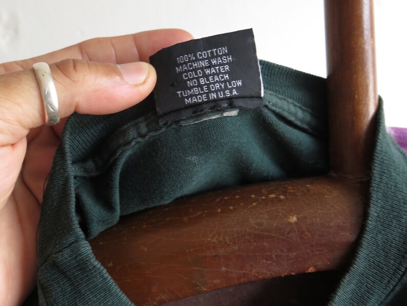 May include: A dark green t-shirt with a black care label that reads "100% Cotton, Machine Wash Cold Water, No Bleach, Tumble Dry Low, Made in U.S.A."