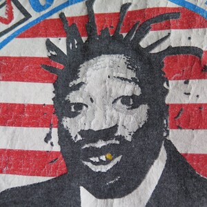 Pu&ograve; includere: Un'immagine in bianco e nero di un uomo con un dente d'oro, che indossa una giacca da abito, con uno sfondo di bandiera americana. L'immagine &egrave; stampata su una maglietta grigia.