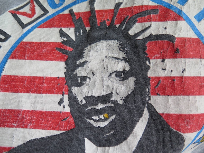 Pu&ograve; includere: Un'immagine in bianco e nero di un uomo con un dente d'oro, che indossa una giacca da abito, con uno sfondo di bandiera americana. L'immagine &egrave; stampata su una maglietta grigia.