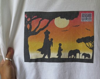 May include: A white t-shirt with a silhouette image of people walking in a sunset landscape. The image includes a tree, a sun, birds, and two elephants. The text "WRM WORLD RELIEF MISSION" is in the top right corner of the image.