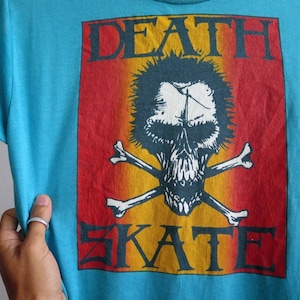 May include: A turquoise blue t-shirt featuring a red, orange, and yellow graphic print of a skull and crossbones with the text "DEATH SKATE".