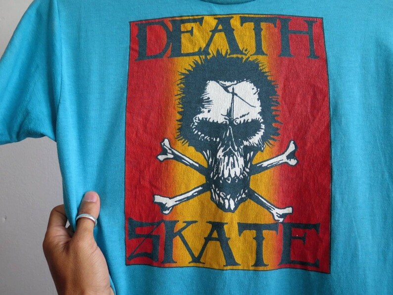 May include: A turquoise blue t-shirt featuring a red, orange, and yellow graphic print of a skull and crossbones with the text "DEATH SKATE".