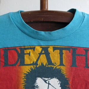 May include: A turquoise blue t-shirt featuring a graphic print of the word "DEATH" in yellow, red, and black lettering. The graphic also includes a black and white image of a person with spiky hair.