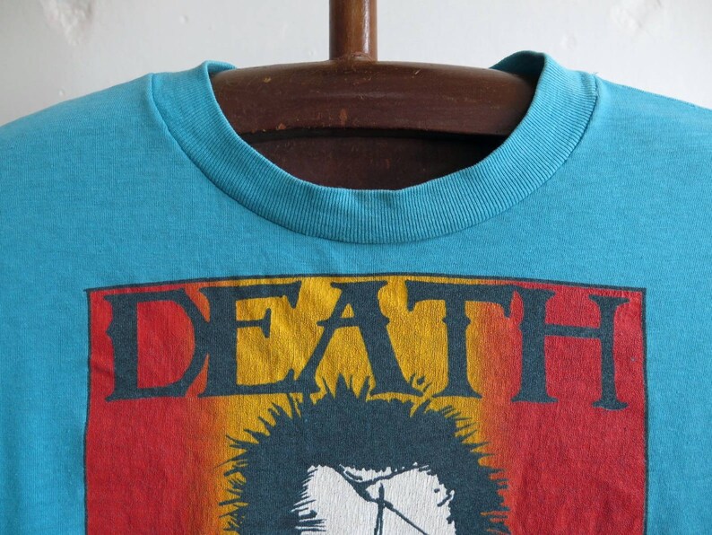 May include: A turquoise blue t-shirt featuring a graphic print of the word "DEATH" in yellow, red, and black lettering. The graphic also includes a black and white image of a person with spiky hair.