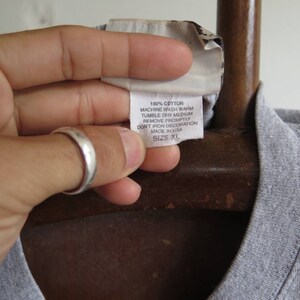 Pu&ograve; includere: Una maglietta grigia con un'etichetta bianca che recita "100% Cotone, Lavaggio in lavatrice a caldo, Asciugatura in asciugatrice a temperatura media, Rimuovere prontamente, Non stirare la decorazione, Prodotto negli Stati Uniti, Taglia XL."