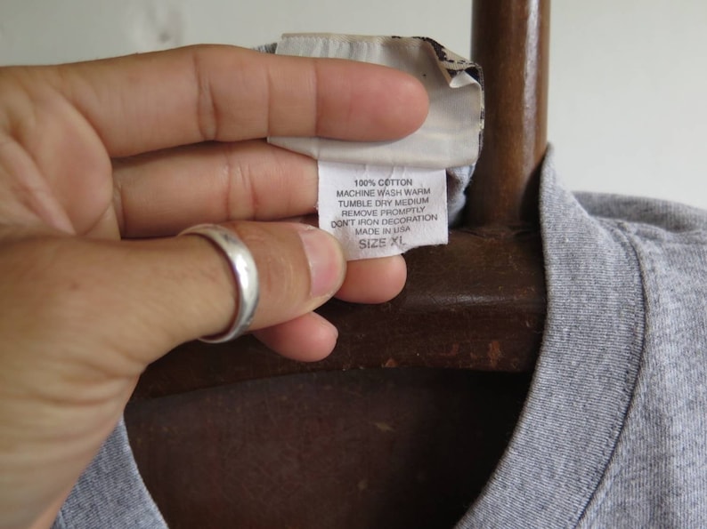 Pu&ograve; includere: Una maglietta grigia con un'etichetta bianca che recita "100% Cotone, Lavaggio in lavatrice a caldo, Asciugatura in asciugatrice a temperatura media, Rimuovere prontamente, Non stirare la decorazione, Prodotto negli Stati Uniti, Taglia XL."
