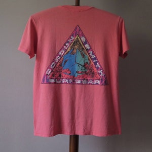 Pu&ograve; includere: T-shirt color corallo con grafica sul retro. La grafica &egrave; un triangolo con le parole "Gordon Smith Surf Gear" e un design colorato con un surfista e un'onda.