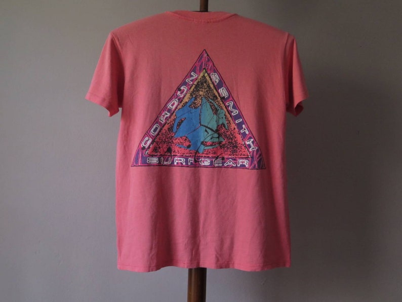 Pu&ograve; includere: T-shirt color corallo con grafica sul retro. La grafica &egrave; un triangolo con le parole "Gordon Smith Surf Gear" e un design colorato con un surfista e un'onda.