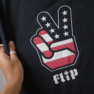 May include: Black t-shirt with a peace sign graphic in the colors of the American flag. The peace sign has the word "FLIP" printed below it.