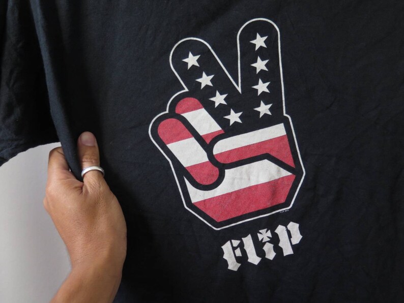 May include: Black t-shirt with a peace sign graphic in the colors of the American flag. The peace sign has the word "FLIP" printed below it.