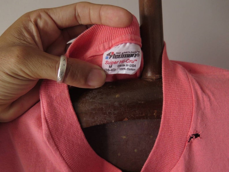 Pu&ograve; includere: Primo piano di una maglietta color corallo con scollo a V. L'etichetta recita "Stedman Super Hi-Cru" e "Made in USA". La maglietta &egrave; taglia M (38-40) ed &egrave; realizzata in 100% cotone. La maglietta ha un piccolo foro vicino allo scollo.