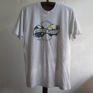 May include: A gray t-shirt with a graphic of two globes with the text "Save the Planet" in a banner between them. The text "One World" is above the globes and "Nature or None" is below the globes.