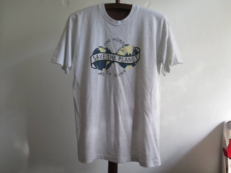 May include: A gray t-shirt with a graphic of two globes with the text "Save the Planet" in a banner between them. The text "One World" is above the globes and "Nature or None" is below the globes.