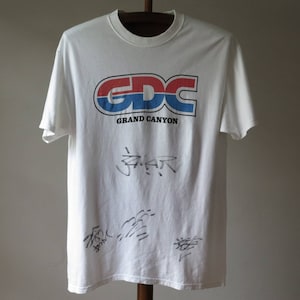 Puede incluir: Camiseta blanca con un logotipo azul, rojo y blanco que dice "GDC GRAND CANYON". La camiseta tiene varias firmas en tinta negra.
