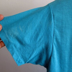 May include: A turquoise blue t-shirt with a red and white design on the right side. The shirt is made of a soft, comfortable fabric.