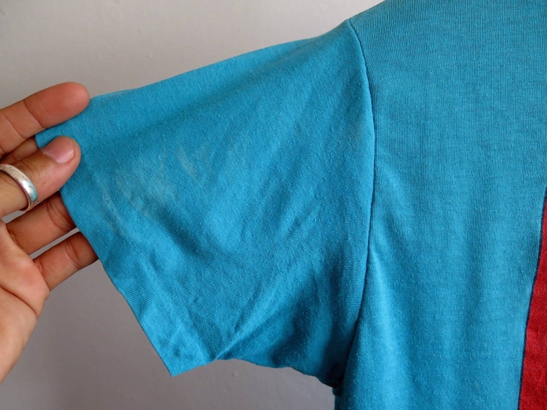 May include: A turquoise blue t-shirt with a red and white design on the right side. The shirt is made of a soft, comfortable fabric.