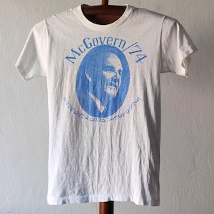 Könnte beinhalten: Weißes T-Shirt mit einem blauen ovalen Grafikdesign, das ein Porträt und den Text "McGovern/74" zeigt. Das T-Shirt enthält auch den Satz "We've got a good thing going" in blauer Schrift. Das T-Shirt wird auf einem Holzkleiderbügel präsentiert.