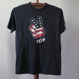 May include: Black t-shirt with a peace sign graphic in red, white, and blue, resembling the American flag. The word "FLIP" is printed below the peace sign.