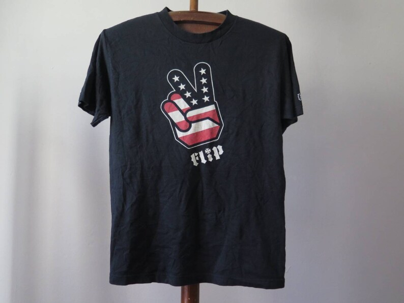 May include: Black t-shirt with a peace sign graphic in red, white, and blue, resembling the American flag. The word "FLIP" is printed below the peace sign.