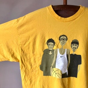 May include: Yellow t-shirt with a graphic print of three cartoon figures. The figures are wearing casual clothing and one is holding a basketball. The text "Nike" is visible on the shirt.
