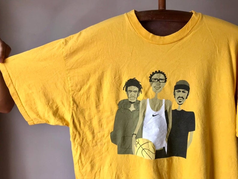 May include: Yellow t-shirt with a graphic print of three cartoon figures. The figures are wearing casual clothing and one is holding a basketball. The text "Nike" is visible on the shirt.