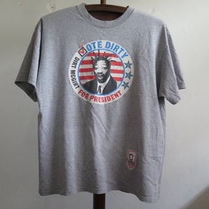 Pu&ograve; includere: Maglietta grigia con una grafica circolare di un uomo con un ciuffo mohawk e il testo "Vote Dirty Dirt McGirt for President".