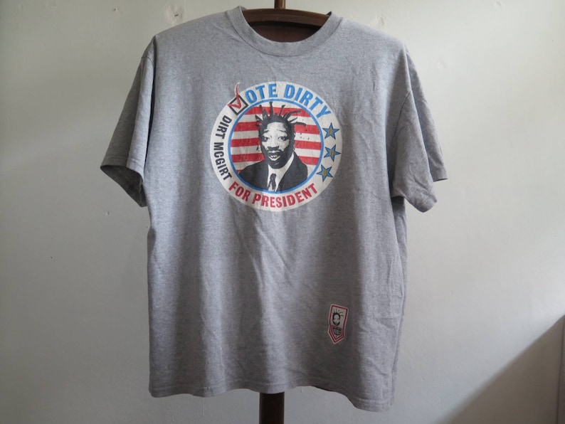 Pu&ograve; includere: Maglietta grigia con una grafica circolare di un uomo con un ciuffo mohawk e il testo "Vote Dirty Dirt McGirt for President".
