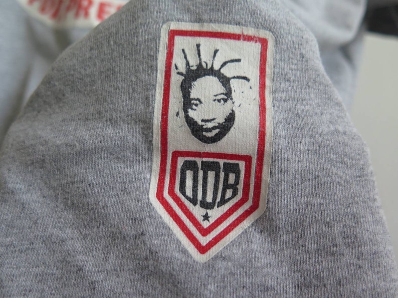 Pu&ograve; includere: Una maglietta grigia con una toppa bianca che presenta un'illustrazione in bianco e nero del viso di un uomo con una stella e il testo "ODB" in nero.