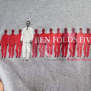 Könnte beinhalten: Ein graues T-Shirt mit einem roten und weißen Grafikdruck einer Band namens "Ben Folds Five". Die Grafik zeigt eine Reihe von Männern in roten Anzügen, wobei ein Mann in einem weißen Anzug in der Mitte steht.