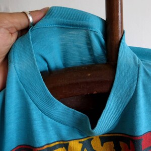 May include: Close-up of a turquoise blue t-shirt with a ribbed neckline. The shirt has a faded look and is hanging on a wooden hanger.