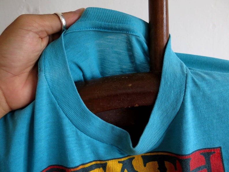 May include: Close-up of a turquoise blue t-shirt with a ribbed neckline. The shirt has a faded look and is hanging on a wooden hanger.