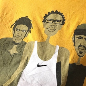 May include: A yellow t-shirt with a graphic print of three cartoon figures. The figures are wearing different clothing, including a tank top with a Nike logo, a hooded sweatshirt, and a t-shirt. The figures are all smiling.