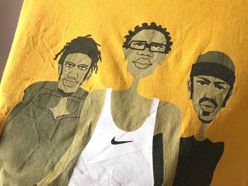 May include: A yellow t-shirt with a graphic print of three cartoon figures. The figures are wearing different clothing, including a tank top with a Nike logo, a hooded sweatshirt, and a t-shirt. The figures are all smiling.