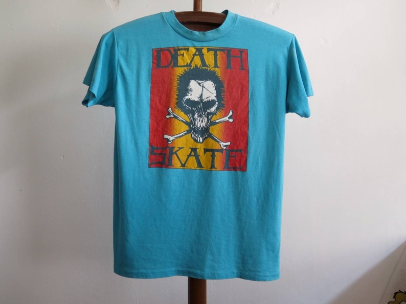 May include: A turquoise blue t-shirt featuring a graphic print of a skull and crossbones. The graphic is in a red, yellow and orange colour scheme. The text "DEATH SKATE" is printed above the skull.