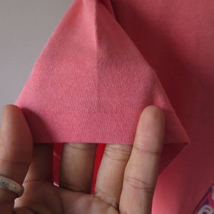 Pu&ograve; includere: Primo piano di una manica di maglietta color corallo. Il tessuto &egrave; una maglia morbida e il bordo &egrave; ripiegato. Il design della maglietta &egrave; parzialmente visibile nell'angolo in basso a destra.