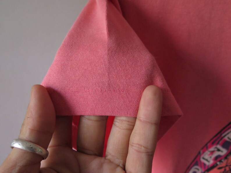 Pu&ograve; includere: Primo piano di una manica di maglietta color corallo. Il tessuto &egrave; una maglia morbida e il bordo &egrave; ripiegato. Il design della maglietta &egrave; parzialmente visibile nell'angolo in basso a destra.