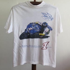 Puede incluir: Camiseta blanca con un gráfico de un piloto de moto en una moto azul y amarilla. El texto "Telefónica Movistar 500cc World Champion" está impreso en la camiseta. El número "1" está impreso en la camiseta con un contorno rojo.