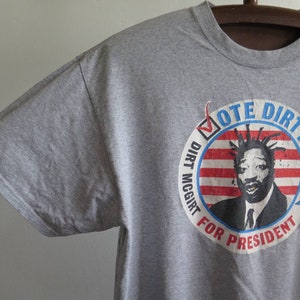 Pu&ograve; includere: Maglietta grigia con una grafica di un uomo con i dreadlocks e un design di bandiera americana rosso, bianco e blu. La grafica dice "Vote Dirty Dirt McGirt For President".