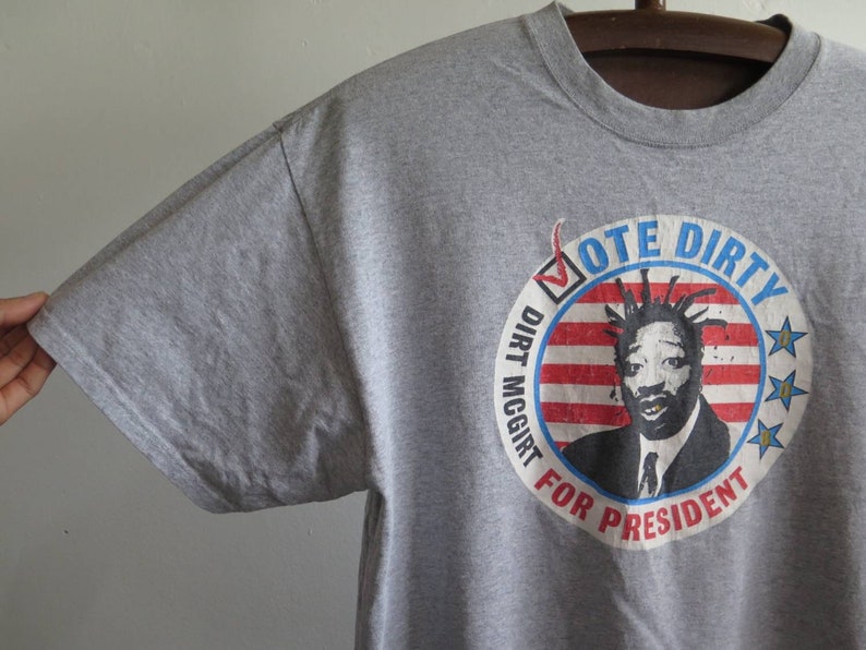 Pu&ograve; includere: Maglietta grigia con una grafica di un uomo con i dreadlocks e un design di bandiera americana rosso, bianco e blu. La grafica dice "Vote Dirty Dirt McGirt For President".