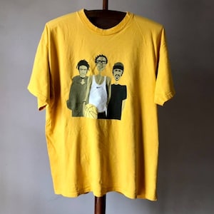 May include: Yellow t-shirt with a graphic print of three cartoon figures holding a basketball. The figures are wearing casual clothing and have exaggerated features.