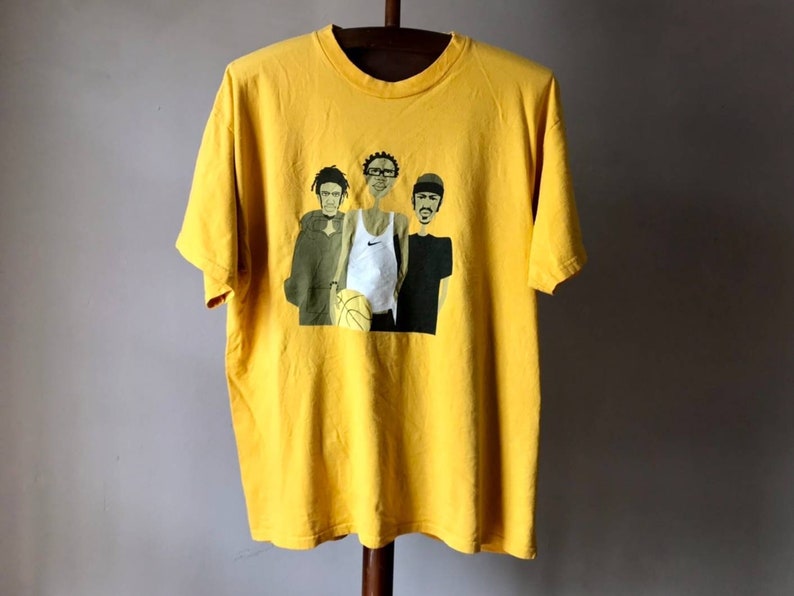 May include: Yellow t-shirt with a graphic print of three cartoon figures holding a basketball. The figures are wearing casual clothing and have exaggerated features.
