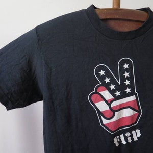 May include: Black t-shirt with a peace sign graphic featuring the American flag colors. The word "Flip" is printed below the graphic.