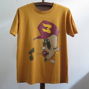 Puede incluir: Una camiseta amarilla mostaza con un personaje de dibujos animados que lleva una gorra de béisbol burdeos y amarilla con la letra "B" en ella. El personaje tiene ojos grandes, un bigote negro y un pendiente de oro.