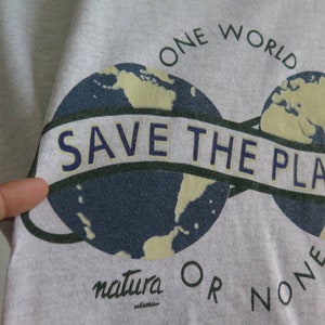 May include: A gray t-shirt with a graphic of two globes connected by a banner that reads "Save the Planet". The text "One World" is above the globes and "natura OR NONE" is below.