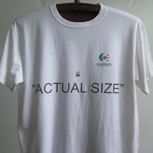 Puede incluir: Camiseta blanca con un logotipo de Logitech y el texto "ACTUAL SIZE" impreso en la parte delantera.
