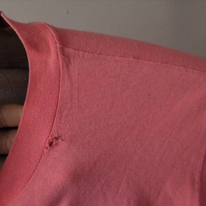Pu&ograve; includere: Primo piano di una maglietta color corallo, che mostra lo scollo e la cucitura della spalla. Il tessuto sembra essere una maglia morbida, con un piccolo foro vicino allo scollo. La manica &egrave; visibile sul lato destro.