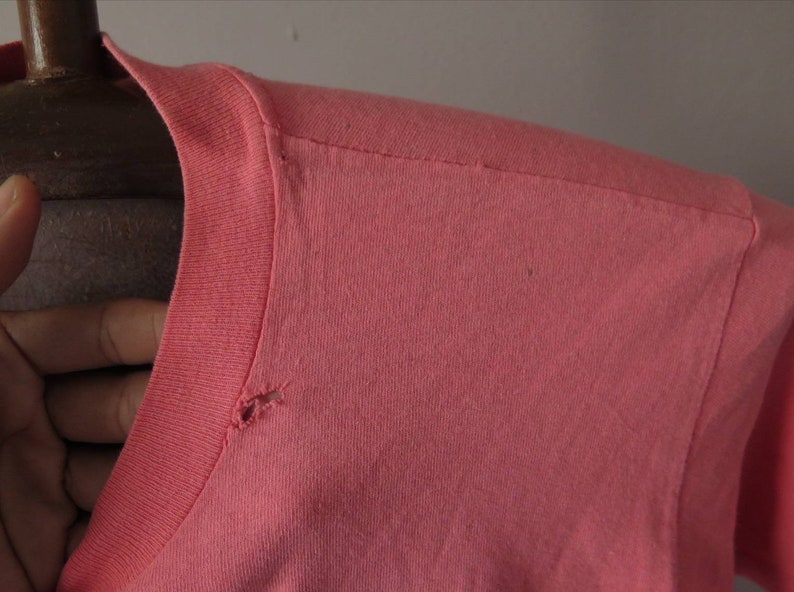 Pu&ograve; includere: Primo piano di una maglietta color corallo, che mostra lo scollo e la cucitura della spalla. Il tessuto sembra essere una maglia morbida, con un piccolo foro vicino allo scollo. La manica &egrave; visibile sul lato destro.