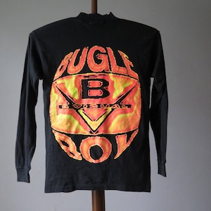 Op de afbeelding: Zwart langarm T-shirt met een cirkelvormige afbeelding van de woorden "Bugle Boy" in een geel en oranje vlammenontwerp.