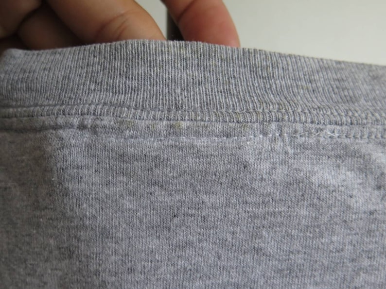 Pu&ograve; includere: Primo piano del collo di una maglietta grigia. Il collo &egrave; a costine e presenta una cucitura doppia.